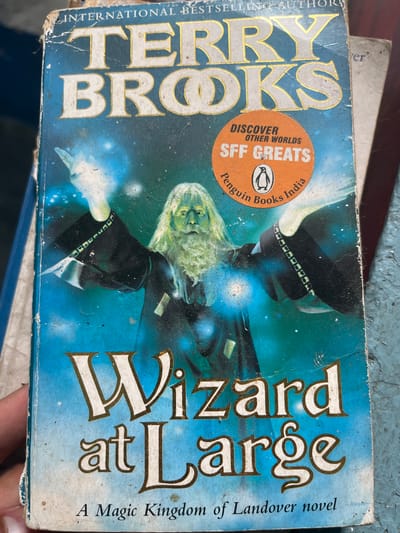 An aged book cover for "Wizard at Large" by Terry Brooks, part of the Magic Kingdom of Landover series, is displayed in Bhagsu, India. The cover features a wizard with a long beard and flowing white hair, clad in a dark robe, with glowing blue energy emanating from his hands. A circular orange sticker with "DISCOVER OTHER WORLDS SFF GREATS Penguin Books India" is affixed to the upper right portion of the cover. The book exhibits wear and tear, with visible creasing and damage to the edges. The background is a vibrant blue, suggesting a magical or celestial setting. The image is taken at a slightly upward angle, with a hand holding the book from the left side. The book is resting on a rough, blue-painted surface.