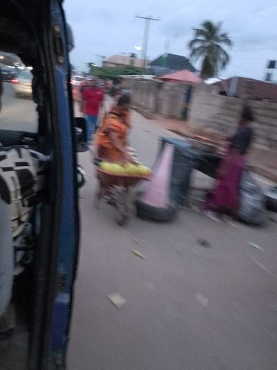 The image depicts a street scene in Orie Uratta, Nigeria, captured from inside a moving vehicle, likely a tricycle (keke napep), indicated by the blue frame and patterned seat visible on the left. The lighting suggests dusk or an overcast day.
In the midground, a woman wearing an orange top and a patterned skirt pushes a brown wheelbarrow loaded with multiple light yellow objects, possibly produce such as plantains or corn, from left to right across the paved road. Further right, another individual, partially obscured and blurred, is seen in a dark top and a magenta skirt, positioned near items on the ground that resemble market goods or discarded materials, including a dark-colored tire and other dark objects.
In the background, multiple blurry figures of pedestrians and vehicles are visible along the street, suggesting ongoing activity and traffic. Buildings line both sides of the road, including structures with a light-colored fence or wall. A tall utility pole with overhead lines is present, alongside a palm tree on the right side of the street. A small structure with a reddish roof is also discernible in the background. The road surface appears grey and scattered with small pieces of debris.