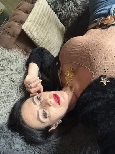 A woman with fair skin, dark hair, and red lipstick is lying on a gray faux fur blanket, likely indoors. She is wearing a light brown lace top, a gold necklace, and a black fuzzy jacket. A decorative gold pin is on the jacket. She is wearing a ring on her left hand, which is near her face. A tufted brown bench is visible, along with a square tan pillow with a striped texture. The location appears to be a private residence. There are no indications of this being in Konz, Germany.