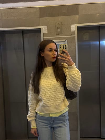 A woman in light blue jeans and a cream-colored knit sweater takes a selfie in front of elevator doors. She holds a cell phone with a patterned case. Her long, dark hair falls down her shoulders. A black purse is visible on her shoulder. The elevator's interior is gray and contains control panels and buttons. The image is vertical and has good lighting.