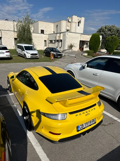 In Arbanassi, Bulgaria, a bright yellow Porsche GT3 is parked in a lot on a sunny day. The car has black racing stripes on its roof and trunk. In the background, a white SUV is parked next to the Porsche. Further back, a white van and a black car are visible, as well as a modern building with a unique architectural design. The ground is asphalt with white parking lines. The sky is blue with scattered clouds.