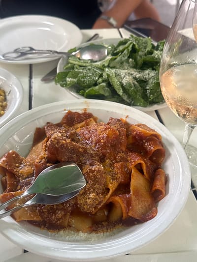 A top-down shot showcases a bright dining table. A glass of white wine is positioned above a small bowl of dark green leafy vegetables. Next to it, a larger oval plate of wide, curled pasta covered in a red sauce and sprinkled with parmesan cheese is presented. Two silver serving spoons are nestled in the pasta. A small white plate and another dish containing yellow food are also visible at the bottom left and bottom center of the image.