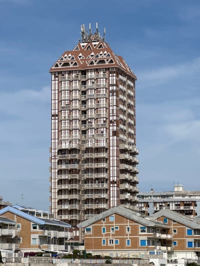 The image showcases the Torre Palazzi, a prominent high-rise building located in Nettuno, Italy. The tower is distinguished by its unique architectural design, featuring a dark brown angular peak adorned with triangular shapes and an array of telecommunications antennas at its apex. The building's facade is characterized by repeating patterns of rectangular windows and balconies, creating a grid-like structure. Adjacent to the tower is a complex of lower-rise buildings of similar style with a terraced design. The sky above is clear and blue, offering a bright background. The photograph seems to have been rotated 90 degrees counter-clockwise.