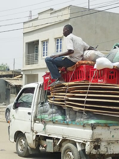 A daytime photograph depicts a white, right-hand drive utility pickup truck, heavily loaded with cargo, on a street in Babban Giji, Nigeria. An adult male, dressed in a light-colored short-sleeved shirt and blue jeans, is seated on top of the cargo bed, facing away from the camera. The truck's cargo comprises a bottom layer of several large, dark-colored sacks secured with a green strap, topped by a substantial stack of flat, oval-shaped wooden or cardboard panels tied with ropes. Above these are multiple red plastic crates, one displaying "Coca-Cola" branding, and various other light-colored sacks and bundled items. In the background, a light-colored two-story building with a flat roof, white-framed windows with light-blue panes, and a second-story balcony with metal railings is visible. To its left, a structure with a corrugated metal roof can be partially observed. Overhead power lines are present, and a silver sedan is partially visible behind the pickup truck.