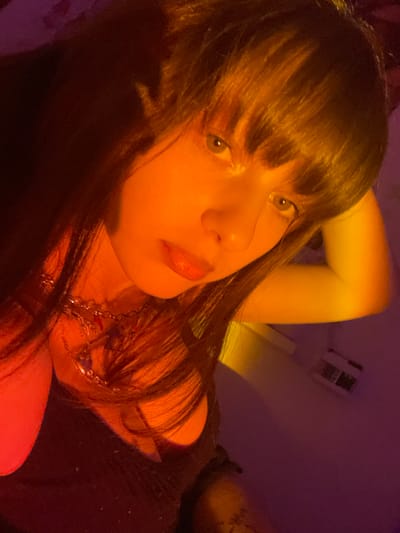 A woman with long brown hair and bangs gazes at the camera with a slightly angled head. She is fair-skinned, with light eyes and slightly parted lips. A silver necklace adorns her neck. The lighting is warm and intense, giving the image a yellowish-orange hue. The background is dark and indistinct, with a few blurred shapes suggesting a room setting. The overall impression is intimate and softly focused.