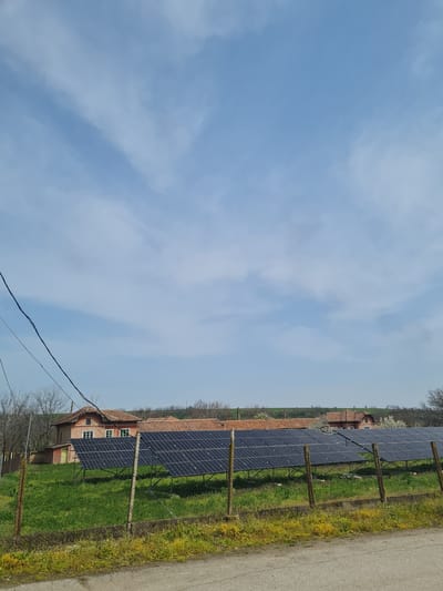 The image depicts a rural scene in Alekovo, Bulgaria. A series of solar panels are erected along a fence line bordering a grassy field. Behind the panels is a red brick building with a red tile roof. A paved road is adjacent to the fence. Overhead power lines run diagonally across the bottom left of the frame. The sky is blue with scattered clouds.
