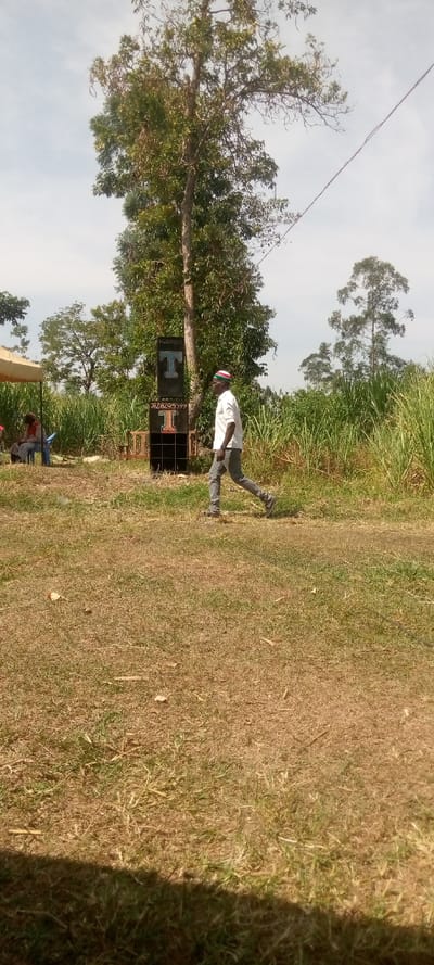The image shows a grassy field in Burundu, Kenya, located at latitude 0.465, longitude 34.74. A man in a white shirt and grey pants, wearing a hat, walks away from the camera toward a large, black, speaker stack. There is a single-person tent in the background, and a woman in a blue shirt and jeans sits next to it. The grass is brown, and the sky is clear. There is a tree behind the speaker stack. The man is walking toward the speaker stack. There is a power line running diagonally across the top of the image. There is a sign on the speaker stack, which is black and has white letters. The sign has two phone numbers on it. The ground is dry and dusty. There are some bushes on the right-hand side of the image. The vegetation is a mix of grass, bushes, and trees. The overall scene is bright and sunny.