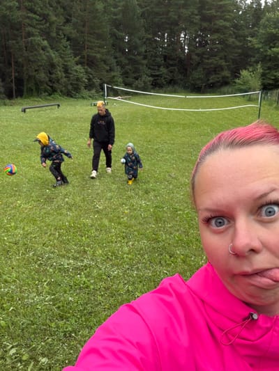 In a grassy, outdoor area with a line of pine trees in the background and a volleyball net stretched across it, a woman with bright pink hair is taking a selfie. Her eyes are wide and her tongue is sticking out playfully, with a nose ring visible. She is wearing a bright pink jacket. In the middle ground, a person in a black hoodie stands, while two children in colorful jackets are playing with a ball in the foreground. One child is actively kicking a colorful ball, and the other is walking with a smaller ball. The overall scene suggests a casual outdoor activity or a park setting. There is no information to suggest a specific location in the city of Прикамский, Russia.
