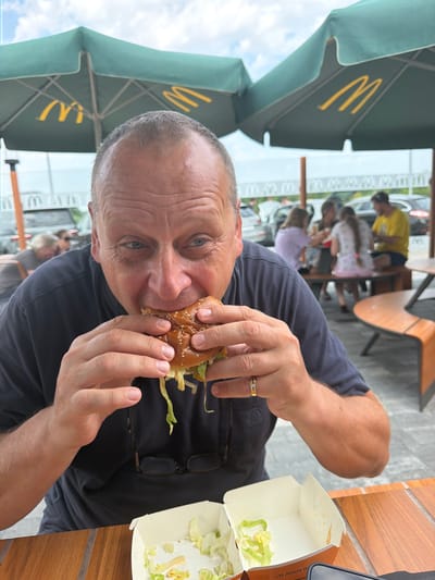 In Budykierz, Poland, a man with short gray hair is captured in a close-up shot, actively eating a hamburger. He is seated outdoors, under a green umbrella with a yellow McDonald's logo. The man is wearing a dark blue t-shirt and a gold ring with a diamond on his left ring finger. His eyeglasses are visible hanging on his shirt. In front of him, on a wooden table, are two white paper food containers, one of which contains lettuce. In the background, other people are visible sitting at tables, and cars are parked in what appears to be a parking lot. The sky is overcast.