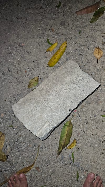 The image displays an outdoor ground scene. The main subjects are a large, light grey, rectangular concrete block or paver, resting on a rough, dark grey ground surface composed of concrete or asphalt mixed with small pebbles and dirt. Scattered around the block are numerous fallen leaves, ranging in color from vibrant yellow and green to dried brown and reddish-brown. A bare human foot, likely an adult's, is partially visible in the top right and bottom right corners, suggesting a person is present, possibly standing or walking.
There is no discernible activity or event taking place; the scene is static. The lighting appears to be low or artificial, possibly indicating dusk, night, or an overcast day, as there are no sharp shadows and colors seem muted. The presence of fallen leaves suggests an outdoor environment with nearby trees, possibly during autumn or a dry period. The location context provided is Cam Đức, Vietnam. No text is visible within the image.