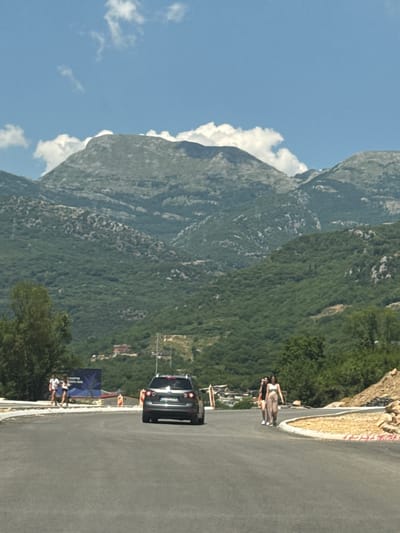 This media file, taken in Prijevor, Montenegro, depicts a scenic mountain landscape under a clear blue sky with scattered white clouds. The dominant feature is a massive, lush green mountain rising steeply, its slopes covered in dense vegetation. A winding asphalt road cuts through the foreground, curving to the right.

Several people are visible along the roadside. Three individuals, two women and a man, are standing near a sign that appears to be a banner for an event or competition. They are dressed casually, suggesting a daytime activity. Further down the road, a black SUV is parked. A group of three women, wearing swimwear, are also present, some appearing to be posing or enjoying the view.

The overall scene suggests a warm, sunny day, likely during the summer months. The location hints at a popular tourist or recreational area, possibly a viewpoint or a place associated with outdoor activities like hiking or driving. The presence of the banner and the people's attire suggest some form of event or gathering is taking place. The visible text on the banner is partially obscured but appears to contain stylized lettering. The natural beauty of the mountains and the blue sky create a serene and picturesque atmosphere.