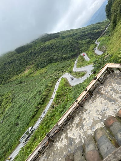 Captured from a high vantage point adjacent to a building with a tile and stone roof, the scene depicts a winding mountain road in Yên Minh Commune, Vietnam. The road traverses a steep, verdant slope, densely covered in vegetation, descending into a foggy valley. Several vehicles, including cars and a truck, are visible on the road, along with a group of cyclists. The road features sharp hairpin turns as it ascends the mountain. The overall perspective offers a panoramic view of the mountainous landscape and the road infrastructure cutting through it.