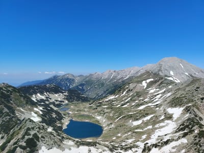 The image captures a panoramic view of a mountainous alpine landscape under a clear blue sky. In the foreground, the terrain is rugged with patches of snow and sparse green vegetation clinging to rocky slopes. Two glacial lakes are visible nestled within the valley; one is larger and deep blue, while the other is smaller and appears lighter blue. Further into the background, a series of imposing mountain ranges stretch towards the horizon, their peaks partly covered in snow. The scene is devoid of any people or man-made structures, presenting a natural and remote environment. This is likely a view of the Pirin Mountains near Bansko, Bulgaria, known for its glacial lakes and alpine terrain.
