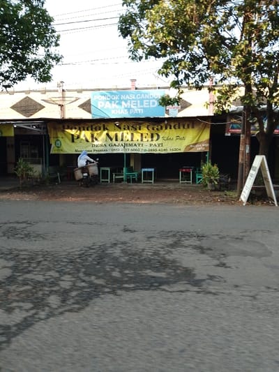 The image shows a street in Pati, Indonesia, with a small restaurant storefront, "Pondok Nasi Gandul Pak Meled," located at coordinates -6.755, 111.038.  A single person, dressed in a white shirt and blue jeans, is riding a scooter in front of the restaurant.  The restaurant is decorated with brightly colored banners, and there is a small sign with the name of the restaurant.  There are small, mismatched tables and chairs set out in front of the building. The street is paved asphalt, with a large puddle in the middle of the frame. The surrounding environment includes a building to the left, lush green trees to the right, and a sign for "Mineral" to the right of the restaurant's sign. The restaurant appears to be open, but the street is mostly empty, with only the motorcyclist and a few chairs and tables.