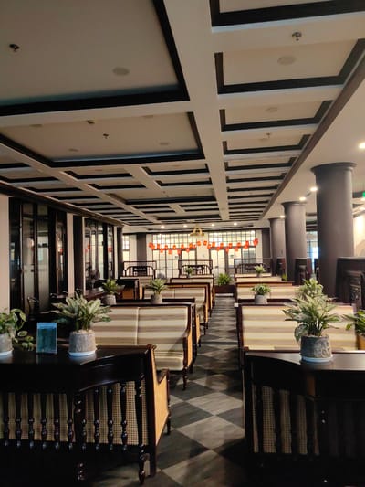 The image captures the interior of an empty restaurant or dining hall, designed with a blend of traditional and modern Asian aesthetics, located in Bai Chay Ward, Vietnam. The scene features rows of neatly arranged dining tables and booths stretching into the background.
The furniture primarily consists of dark wooden tables with intricate designs, featuring latticework or cane detailing on their sides. Matching dark wooden chairs, many with cane backs and sides, accompany these tables. The booths are upholstered with light-colored, vertically striped fabric in cream and beige tones, suggesting comfort. Potted green plants, possibly Aglaonema, are placed as decor on each table and along ledges, adding a touch of nature to the space.
The floor exhibits a striking dark checkered pattern, contrasting with the lighter ceiling. The ceiling itself is a prominent feature, showcasing an elaborate coffered design with recessed lighting fixtures embedded within white rectangular panels framed by dark brown or black trim. Tall, dark cylindrical columns line the right side of the hall, while the left side features large glass partitions or windows.
In the background, beyond the dining area, red banners or lanterns are strung horizontally, suggesting a festive or traditional ambiance. The artificial lighting from the ceiling illuminates the space, indicating that it is well-lit, though the time of day is not explicitly discernible.
No people are present, giving the impression that the restaurant is either closed, awaiting customers, or captured during an off-peak hour. On a menu or sign visible on a table in the foreground on the left, the word "DANSOON" is clearly legible. The overall atmosphere is calm, orderly, and ready for service.