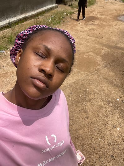 A young Black female with her eyes closed, wearing a pink bonnet with a floral pattern and a pink t-shirt with white lettering, is pictured in a close-up selfie. The t-shirt has the word "Balenciaga" printed on it, with two stylized characters above it. A small wad of Nigerian Naira currency, with "200" visible on the bills, is tucked into the bottom edge of the t-shirt. The background consists of a dirt path with scattered debris and vegetation. In the upper background, a person in dark pants and white sandals walks away from the camera. The overall setting appears to be outdoors in a rural or semi-urban area, possibly on a street or path in Rafin Par, Nigeria. The lighting suggests it is daytime.