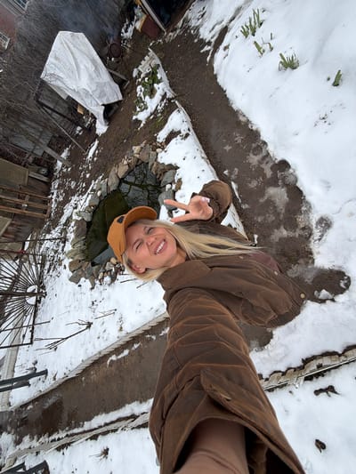 A blonde woman in a brown jacket and cap is taking a selfie in a snowy outdoor location in Sarapul, Russia. She is holding up a peace sign with her left hand. Behind her, there is a small pond surrounded by stones. The ground is covered with snow, and there are various objects visible, including a white tarp, a metal structure, and some plants. The picture is taken from a high angle.