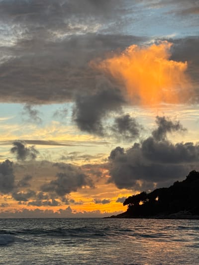 This is a vertical, eye-level, medium shot of a dramatic sunset over the ocean. The sky is filled with a mix of dark, stormy clouds and vibrant streaks of orange and yellow light. The sun appears to be setting behind a dark silhouette of a cliff face on the right side of the image. The ocean, on the far right, reflects the sunset's colors and has a rippled texture, suggesting moderate waves. The scene conveys a sense of awe and the powerful beauty of nature. The time of day is clearly sunset, and the weather appears to be transitioning from stormy to clear, with dramatic cloud formations. The location is identified as Karon, Thailand, which is consistent with a tropical sunset over the ocean. There is no visible text in the image.