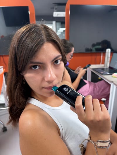 A young woman with brown hair and brown eyes is holding a black Waka vape pen with a blue tip to her lips. She is wearing a white tank top and a bracelet on her right wrist. She is indoors in a room with orange and grey walls, a monitor, and a desk with a laptop on it. Another woman is sitting in the background. The location is San Andrés Cholula, Mexico.
