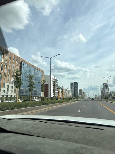 This is a vertical, eye-level, outdoor shot taken from the perspective of a vehicle moving along a multi-lane road. The road has a yellow dividing line and white lane markings. On the left side of the road are several modern, multi-story buildings with glass facades and balconies, interspersed with trees and streetlights. On the right side, a portion of the vehicle's interior, possibly a dashboard or door panel, is visible, covered in a textured gray material. The sky is cloudy with patches of blue, suggesting a partly cloudy day. The scene appears to be a contemporary urban environment, likely in Astana, Kazakhstan, given the context. The time of day appears to be daytime, likely late morning or early afternoon, given the bright, diffused light. There are no visible people in the frame. The overall impression is one of urban development and modern architecture under a dynamic sky.