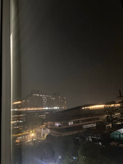 The image depicts a nighttime view of a city, likely in Central Jakarta, Indonesia, given the contextual information. The scene is characterized by a dark, hazy atmosphere, suggesting fog or heavy smog, which obscures distant details and lends a somber mood to the cityscape.
Dominating the right side of the frame are several modern buildings. One prominent, tall building is illuminated with numerous windows, each glowing with a warm, artificial light, indicating occupancy or internal lighting. The side of this building also features a striking vertical strip of bright, yellow lights, possibly accentuating its architectural design or serving as an advertisement. Adjacent to it, or perhaps part of the same complex, is another building with a more distinct facade, revealing balconies and internal structures. The lights within these buildings create a strong contrast against the dark sky.
Trees with dark foliage are interspersed between the buildings, their forms softened by the dim light and atmospheric conditions. The overall impression is one of a dense urban environment at night, where the artificial glow of human habitation battles with the natural darkness and atmospheric obscurity.
In the foreground, the bottom of the image reveals what appears to be a windowsill or a similar interior ledge, suggesting the photograph was taken from inside a building, looking out. The presence of this foreground element grounds the image and reinforces the perspective of an observer within the city.
There is no visible text in the image. The time of day is clearly night, and the weather is characterized by low visibility due to haze or fog. The emotions evoked are likely a mix of awe at the urban landscape and perhaps a sense of isolation or mystery due to the darkness and obscured visibility.