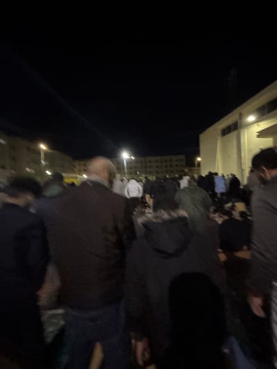From a low angle at night, numerous people gather near a building lit by dim, warm lights. Clothes, possibly belongings, are scattered amongst the crowd. Individuals wearing dark clothing appear to be the focus of the scene, their features indistinct due to low lighting. The overall atmosphere suggests a group experiencing adversity or seeking refuge, with an underlying sense of chaos and uncertainty.