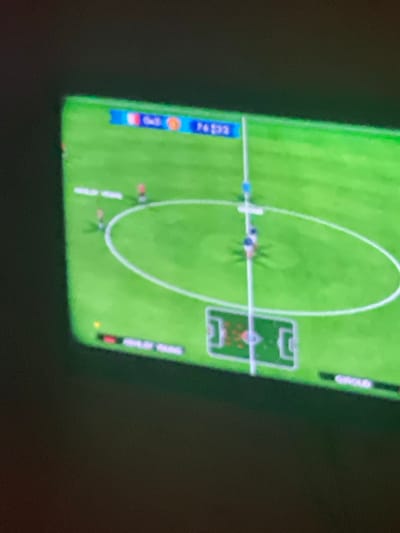 A soccer game is shown from a top-down perspective. A bright green field is marked with white lines, including a center circle and goal areas. Several miniature figures, presumably representing soccer players, are scattered across the field. A white ball is positioned in the center circle. Scores and team colors are visible along the left and right edges of the screen.