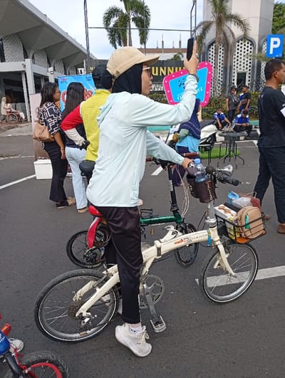 A person on a cream-colored folding bike holds up a phone, seemingly taking a picture. They wear a tan baseball cap, hijab, glasses, light blue jacket, and black pants with white shoes. The bike has a front basket filled with items. Behind them are several other people, including someone in a yellow shirt, and a parked green folding bike. In the background are palm trees and a building. A blue sign with pink trim is visible to the right of the person on the bike.