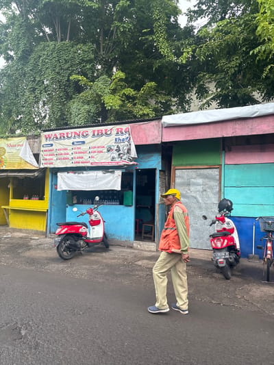 A man in a yellow hard hat and orange vest stands near a colorful roadside shop called "Warung Ibu Ra," offering items like coffee, tea, and juice. The shop is painted with various bright colors and banners advertising its offerings. Two parked red and white motorbikes sit near the entrance, with one possibly obscuring part of the shop. Trees provide shade along the side of the street.