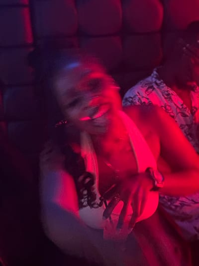 This is a blurry, low-light image with prominent red and blue lighting, suggesting a nightclub or bar setting in Howayi, Nigeria. The primary subject is a person, their face and body significantly blurred due to motion, making detailed identification impossible. The person appears to be wearing a white or light-colored top and patterned bottoms. Their right arm is visible, resting on a dark, possibly leather, surface, possibly a couch or booth. A watch is visible on their wrist. The overall impression is of a candid, possibly candid shot taken during a moment of movement or excitement. The lighting creates a vibrant and somewhat disorienting atmosphere. No text is visible in the image.