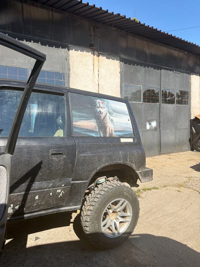 An outdoor, eye-level, medium shot captures a black, dusty, four-door SUV parked on a gravel and concrete surface in front of a large industrial building. The building features a corrugated metal roof and large, dark metal double doors with glass pane inserts. The SUV has its driver's side door open and a sticker of a wolf on the rear passenger window. The environment appears to be a rural or industrial area, possibly on the outskirts of the city of Haskovo, Bulgaria, given the overall aesthetic and natural lighting conditions. No people are visible in the frame, and the scene appears static.