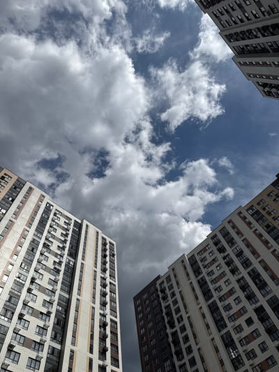 This is a low-angle, exterior, daytime shot of three modern, multi-story residential buildings with a visible sky. The buildings are predominantly light-colored with horizontal bands of darker color and rows of windows. Some windows appear to be open. The sky is a mix of blue and white clouds, with some darker, more ominous clouds present. The buildings appear to be situated in a way that they form a frame around the sky. There are no visible people, text, or specific location cues other than the general context of modern residential architecture. The overall mood is neutral, depicting an ordinary day with a dramatic sky.