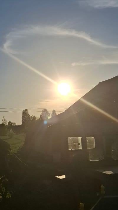 The image shows a scene with a bright sun in the sky. There are thin clouds visible. To the right of the sun, there appears to be a building or structure, possibly with windows. Trees and greenery are visible along the bottom edge of the image. There is no evidence to suggest it is in the city of Шешурга, Russia. The image primarily depicts natural elements (sky, sun, clouds, trees) and a structure, possibly a building.