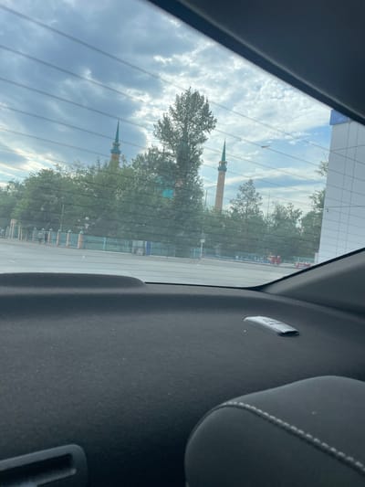 The image, taken from inside a vehicle, shows a view through the rear window of a city street scene. Two prominent mosque minarets with green conical tops are visible above a line of trees. The minarets are cylindrical and appear to be constructed of brick or stone. A modern building with a white tiled facade is to the right of the frame. In the foreground, the dark, textured surface of the car's interior, including part of a seat, is visible. A small, metallic object, possibly a remote or tag, rests on the car's dashboard. The street appears to be an urban road with a sidewalk and a fence along the side, suggesting a public area. The sky is partly cloudy, with a mix of blue sky and gray clouds.
The mosque minarets are recognizable as belonging to the Masgut-Ata Mosque in Pavlodar, Kazakhstan, due to their distinctive architectural style and relative location to other landmarks in the city. The modern building on the right is likely a commercial or administrative building in the vicinity of the mosque. The overall scene depicts a typical urban environment in Pavlodar, Kazakhstan, on what appears to be an overcast day.