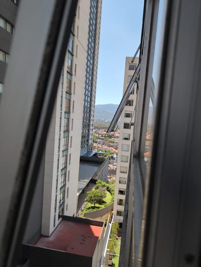 View from an open window in Jesús del Monte, Mexico. Two tall, modern, white buildings with rows of windows dominate the view, separated by a green area and a lower building with a red roof. In the distance, a mountain range is visible above the red-tiled roofs of a residential area. The window frame partially obscures the scene.