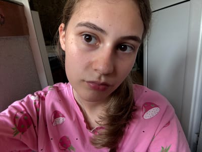 A young woman with light brown hair and brown eyes is shown in a close-up, selfie-style shot. She is wearing a pink pajama top adorned with a pattern of strawberries and dragon fruit. Her hair is pulled back into a ponytail and cascades over her right shoulder. The background is softly blurred, showing elements of a room, including a white doorframe and a dark, textured surface on the left. The lighting appears to be natural, possibly from a window. The image was taken indoors. There is no information available about the specific location in the city of Panagyurishte, Bulgaria.