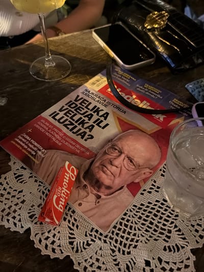 The image depicts a close-up, overhead view of a table setting in what appears to be a bar or cafe, possibly in Belgrade, Serbia, given the Cyrillic text on the magazine. A glass with a yellow beverage and ice sits on the left, partially out of frame. In the center, a magazine is spread open, its cover featuring a portrait of an elderly man with glasses. The magazine's title is in Cyrillic and appears to be "Nedeljnik". Below the man's portrait, there is text that includes "MILAN ST. PROTIĆ", "SEDAM DECENIJA", and "VELIKA IDEJA VELIKA ILUZIJA?". A red packet of "Smoking RED" cigarettes lies diagonally across the magazine. To the right, a glass of water with ice is visible, and further to the right, a black clutch purse with a gold clasp and a smartphone are placed on the table. The table surface is dark wood, and the items are arranged on a white, crocheted doily. The background is dark and slightly blurred, suggesting an indoor, dimly lit environment.