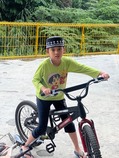 A young male, appearing approximately 7-9 years old, stands centrally positioned, straddling a black BMX-style bicycle on a light gray concrete surface. He wears a black peci (songkok) adorned with white geometric embroidery, a light green long-sleeved shirt featuring a multi-color cartoon character graphic and text, dark blue cropped pants, and blue flip-flops. His hands grip the black handlebars of the bicycle, which has a red front fork and knobby tires. The bicycle frame prominently displays the word "PATRIOT" in white lettering. The subject's gaze is directed forward, towards the viewer. In the background, a bright yellow painted metal fence runs horizontally, behind which dense green foliage from various plants is visible. A portion of a smaller, light-colored object, possibly another bicycle part, is visible on the ground to the lower left. The scene depicts an outdoor setting, consistent with a residential or recreational area within a city such as Kecamatan Pati, Indonesia.