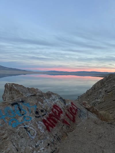 A landscape view presents a distant horizon line amidst a cloudy sky, reflecting on a flat surface resembling water or a salt flat. Close up, a rough rock formation covered in graffiti anchors the foreground. The graffiti includes red and blue lettering, contrasting with the muted tones of the natural setting.