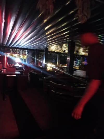 The image depicts the interior of an establishment, possibly a bar or restaurant, with a corrugated metal ceiling illuminated by various colored lights, including red and yellow. The lighting creates a dramatic atmosphere, with beams of light visible. The space appears to be furnished with tables and seating, suggesting it is a place for patrons. In the foreground, a person's arm and shoulder are blurred due to motion, illuminated by red light. The background shows more of the interior, including structures that could be pillars or bar areas, and more seating arrangements. The overall scene is dimly lit, characteristic of a nightlife venue. The image does not provide any specific indicators of its location in Howayi, Nigeria.