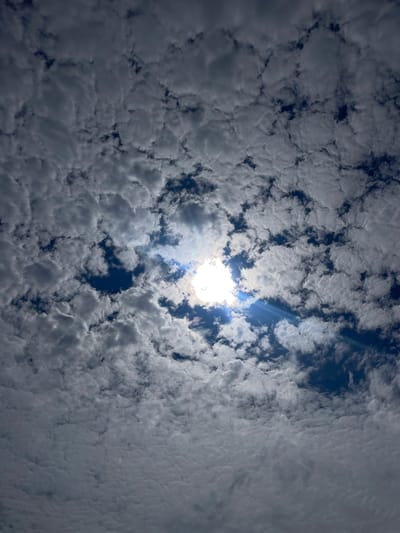 The image shows a sky filled with fluffy, white cumulus clouds interspersed with patches of deep blue sky. The sun is visible, partially obscured by clouds, creating a bright, glowing effect with visible light rays. The clouds appear to be arranged in a somewhat textured, almost rippled pattern across the frame. There are no people, structures, or discernible landmarks visible in the image, suggesting it is a wide-angle shot focused solely on the sky. The scene is devoid of any specific location markers that would pinpoint it to Diepenbeek, Belgium.