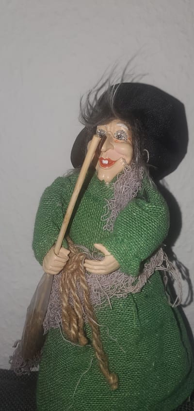 A close-up, eye-level, medium shot captures a doll representing a witch. The doll has a light-colored face with a large, pointed nose, rosy cheeks, a wide red smile, and exaggerated eyelashes decorated with glitter. Its grey hair is wispy and appears windblown. It wears a black, wide-brimmed hat and a coarse, green woven dress. A light purple, frayed scarf is draped around its neck. The doll holds a light-brown wooden stick in its right hand, with a thick, braided brown rope wrapped around its middle and the stick. Its left hand is slightly open, positioned near the rope. The background is a plain, textured light gray wall, with a shadow cast by the doll on the right side. The doll is positioned indoors, and the scene is static. The specific location in the city of Sasbach, Germany is not discernible from the image.
