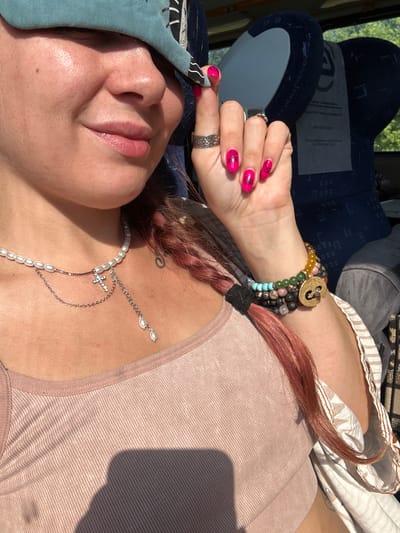 A woman is seated on a bus, with a turquoise eye mask pushed up onto her forehead. Her face is partially obscured, but she appears to be smiling. She is wearing a light pink tank top and a pearl necklace with a cross charm. She also has several bracelets on her wrist, including one with a gold charm. Her fingernails are painted bright pink and decorated with black eye designs. Her hair is a reddish-brown color and is braided. The background shows the interior of a bus, with blue seats visible. The image is taken in natural light, likely during the day. There is no information available to indicate the location within the city of Pokrov, Russia.