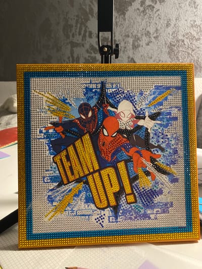 The image shows a square, framed artwork featuring Spider-Man, Miles Morales, and Ghost-Spider. The artwork is adorned with numerous small, sparkling rhinestones in gold, blue, silver, red, and black. The characters are depicted in dynamic poses, with action lines and a distressed brick wall background. The phrase "TEAM UP!" is prominently displayed in large, yellow, rhinestone-covered letters. The frame itself is also decorated with multiple layers of gold and blue rhinestones. The artwork is displayed on a stand, and in the background, a textured gray wall is visible. The scene is set indoors, on a white surface that appears to be a desk or table, with some papers and a pencil visible at the bottom of the frame. There is no discernible connection to Bietigheim-Bissingen, Germany.