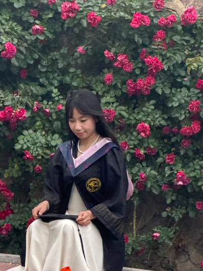 A young East Asian woman wearing a graduation gown and cap is seated in front of a dense wall of pink roses. Her gown is black with purple and pink trim around the neckline and embroidered gold trim on the sleeves. She is holding her graduation cap in her lap. She has on a pearl necklace. The background is filled with lush green leaves and many pink roses. The lighting suggests it is daytime.
