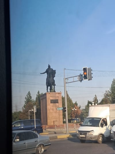 The media file shows a street scene in Shymkent, Kazakhstan. The image is oriented vertically, with the sky occupying the left side and the street and its surroundings on the right. A traffic light is visible, displaying an orange signal. In the background, a statue of a person on a horse stands prominently against the sky, mounted on a stone pedestal.

Several vehicles are present, including a white truck with its headlights on, a blue van, and a silver car. The truck appears to be a commercial vehicle, possibly a cargo van, with a distinctive design. The silver car is parked or stopped on the right side of the street.

The setting appears to be a city street with trees lining the area. The time of day seems to be late afternoon or early evening, given the quality of the light. The sky is clear and blue. There is a sense of everyday urban activity.

On the stone pedestal of the statue, there is a plaque with some text, but it is not legible due to the image quality and distance. The traffic light has a sign attached to its pole with text that is also too small to read. No explicit emotions are conveyed.