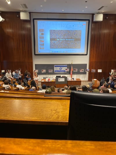 The image depicts a large lecture hall within a building at Princeton University, Princeton, United States. The hall features high, light-colored wood-paneled walls and tiered wooden seating arrangements. Approximately 30-40 individuals, predominantly young adults, are present in the room. A group of individuals is standing and interacting on the left side of the hall, near the front, while others are seated in the wooden benches, generally facing the stage area.
At the front of the hall, a large projector screen displays a PowerPoint slide. The slide prominently features the text "In their memory" above a grid or mosaic of numerous small images. Below the screen, a dark wooden podium, displaying the Princeton University shield logo, is positioned on a stage. Behind the podium, an Israeli flag and an American flag stand upright on poles. Several display easels with printed materials, consistent with photographs or informational posters, are arranged across the stage and along the back wall, suggesting an exhibition or memorial display. White speaker units are mounted high on the walls. The scene indicates an academic event or memorial gathering, either in progress or recently concluded.