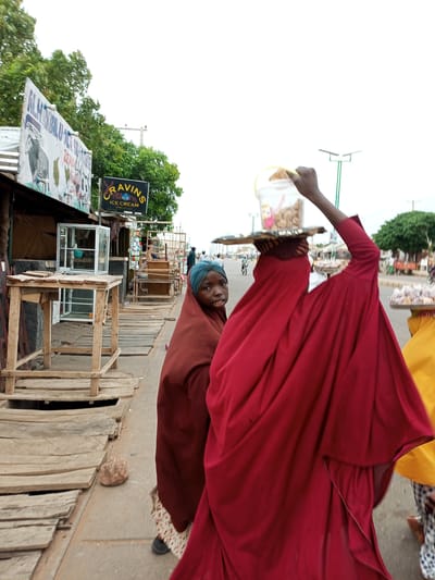 In Tempure, Nigeria, two young females are depicted on a sidewalk. The girl on the left, wearing a brown headscarf and brown dress, is looking directly at the camera with a slightly open mouth. To her right, a second girl, covered in a red cloak with a blue headscarf, is carrying a tray with a bucket of food on her head, holding it with her right hand. The scene is set on a street with a wooden sidewalk made of planks. In the background, there are storefronts with signs, including one that reads "CRAVINS ICE CREAM." Other people and a motorcycle are visible further down the street, and trees line the roadside. The sky is overcast.
