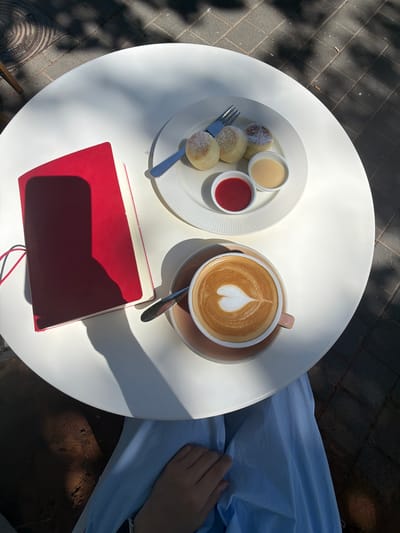 The scene depicts an outdoor cafe setting in Bat Yam, Israel, on a sunny day with strong directional light creating prominent shadows. A person is seated at a white, circular table, with only their lower body and right hand visible. They are wearing a light blue garment, possibly a dress or skirt, with their hand resting on their lap.
On the table, positioned from top to bottom:
1. A white ceramic plate with a fluted rim holds three small, round, baked items, resembling syrniki or small pancakes, dusted with powdered sugar. A small, light blue-handled fork is placed on the plate.
2. Adjacent to the plate, two small white ramekins contain sauces: one with a vibrant red, thick liquid (likely a berry jam), and the other with a pale yellow, creamy liquid (possibly condensed milk or custard).
3. A light pink/beige ceramic coffee cup with a matching saucer holds a beverage featuring distinct heart-shaped latte art. A silver spoon rests beside the cup.
4. To the left, a red hardcover notebook or journal is visible, partially open, with a red elastic band. A rectangular shadow, consistent with a mobile phone, is cast across its cover.
The ground surrounding the table is paved with dark grey bricks, and overhead foliage casts dappled shadows across the scene.
