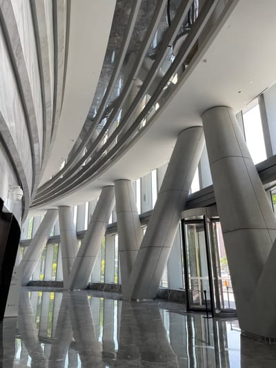 This image shows the interior of a modern building in Chaoyang District, China. The architectural design features a combination of geometric shapes and natural light. Round, gray structural beams intersect with white walls and ceilings. A series of curved, stepped platforms with a marble-like finish are visible. Reflections of the building's exterior and interior are cast onto a glossy surface, possibly a wall or floor. The overall color palette is primarily gray, white, and silver, with hints of green from the outside vegetation. A black frame structure with glass panes is also visible.